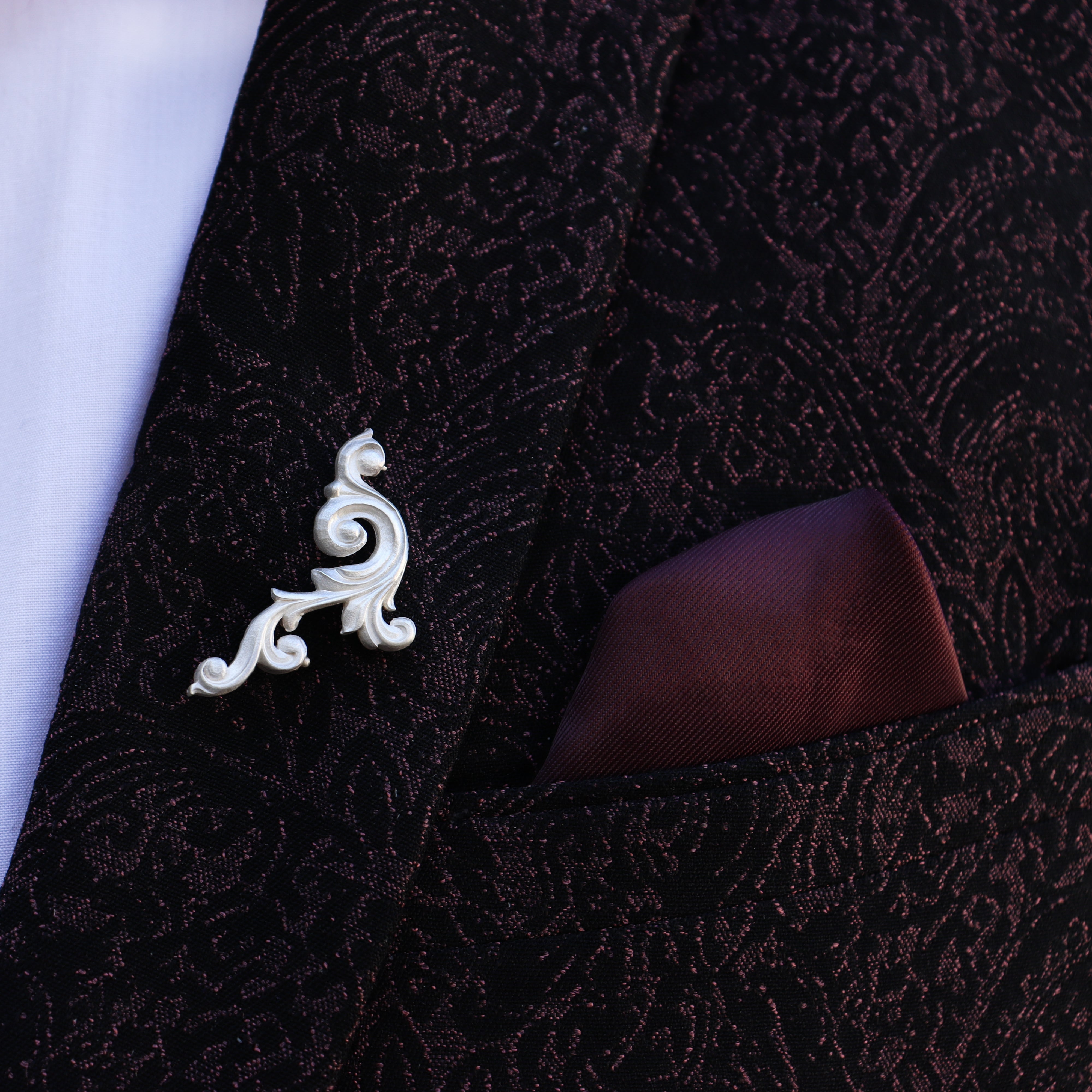 Close-up of a decorative  scroll lapel pin on a dark textured suit jacket.